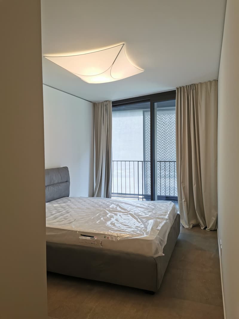 room with a bed, plastic covered mattress, white walls, ceiling light, balcony with a black railing