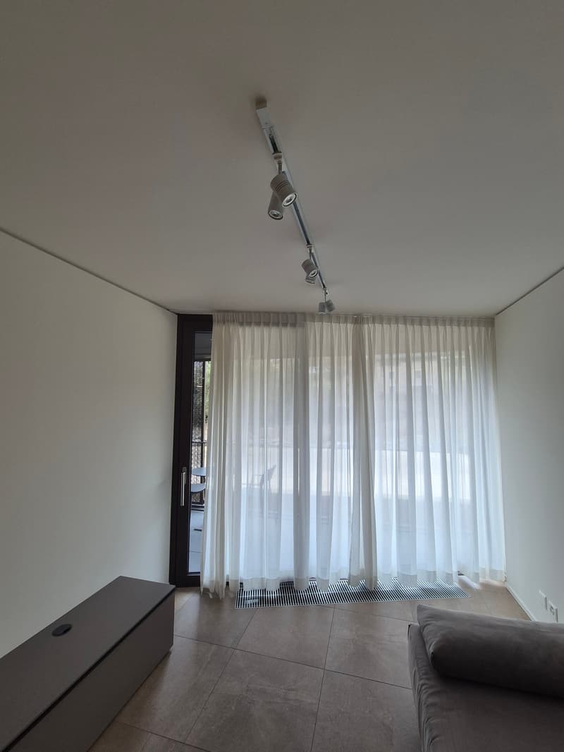 white walls, ceiling with mounted lights, white curtains covering sliding glass door to balcony, large rectangular sofa, ottoman, tile floor