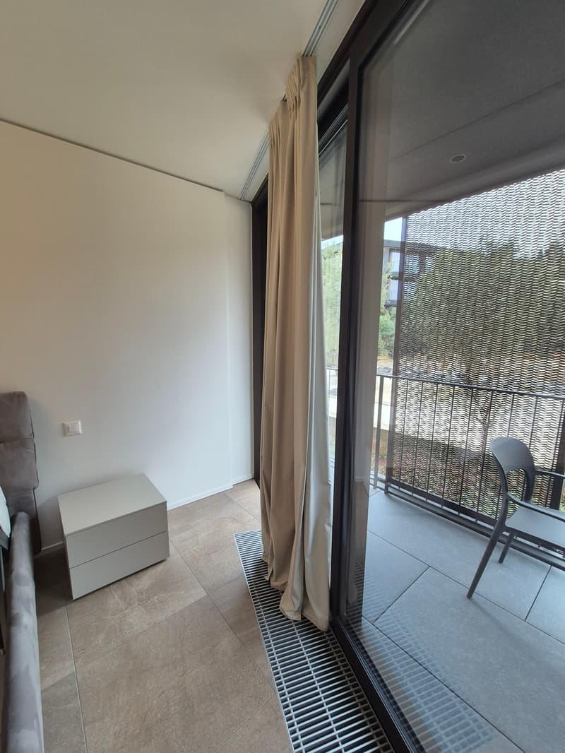 tiled floor, white nightstand, white sliding glass door, beige curtains, balcony with black railings, outdoor chair