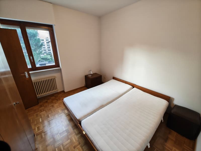 Two single beds, wooden floor, white walls, window with view, nightstand, radiator, lamp, stool