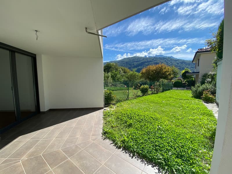 A view of a balcony with tile flooring, overlooking a well-manicured lawn with plants and trees. The sky is blue with clouds.