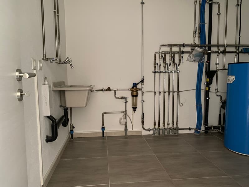 A utility room with white tiled floor, sink, pipes and plumbing fixtures mounted on the wall