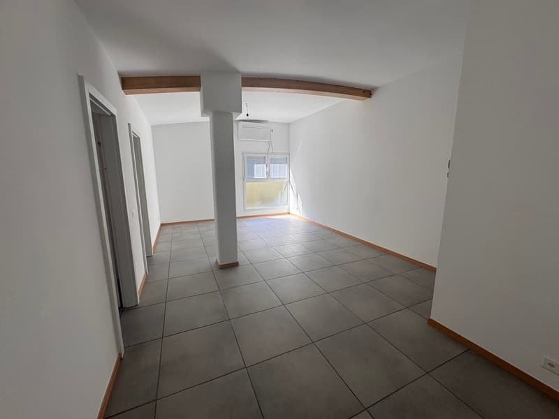 Empty room with white walls, gray tiled floor, wooden beam on ceiling, window with blinds, and an air conditioning unit.
