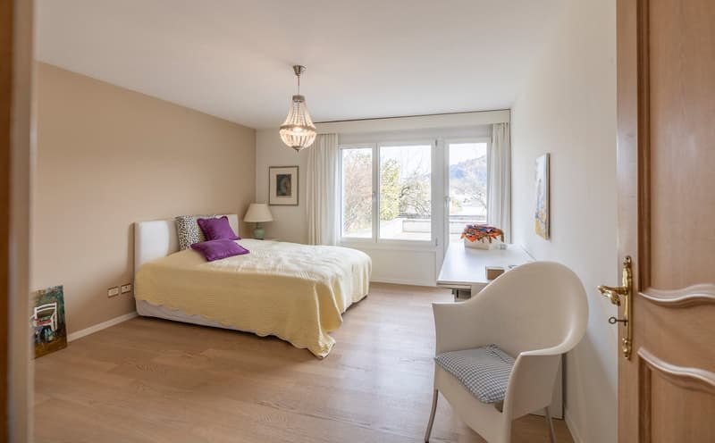 A bedroom with a bed, lamp, nightstand, and a chair. The bed has a blanket and pillows. There is a painting on the wall and a desk with a box and a vase. The room has a chandelier and a window with curtains.