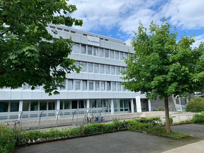 A multi-story office building with a large sign that says 'GLANDON' on the facade. The building has many windows and is surrounded by trees and greenery.