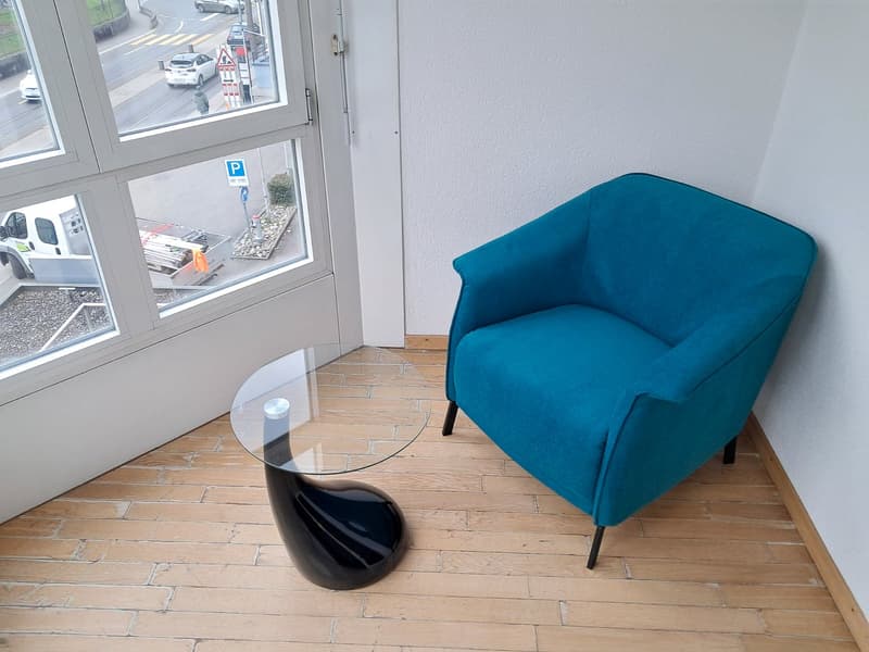 The image shows a small office or commercial space with a large window overlooking a parking area. There is a teal-colored armchair and a glass-topped coffee table with a black base in the foreground. The floor is tiled with a stone-like pattern.