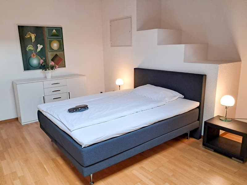 The image shows a bedroom with a double bed, a dresser, and a side table. The room has white walls, hardwood floors, and is well-lit with lamps. There is a framed artwork on the wall above the dresser.