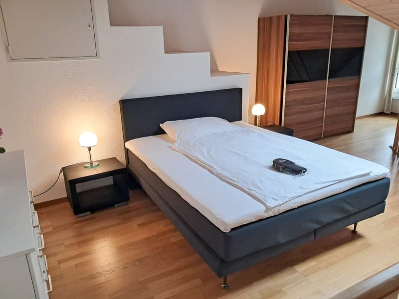 Spacious bedroom with a king-size bed, dark gray headboard, wooden wardrobe, and bedside tables with lamps. The room has hardwood flooring and a modern, minimalist design.