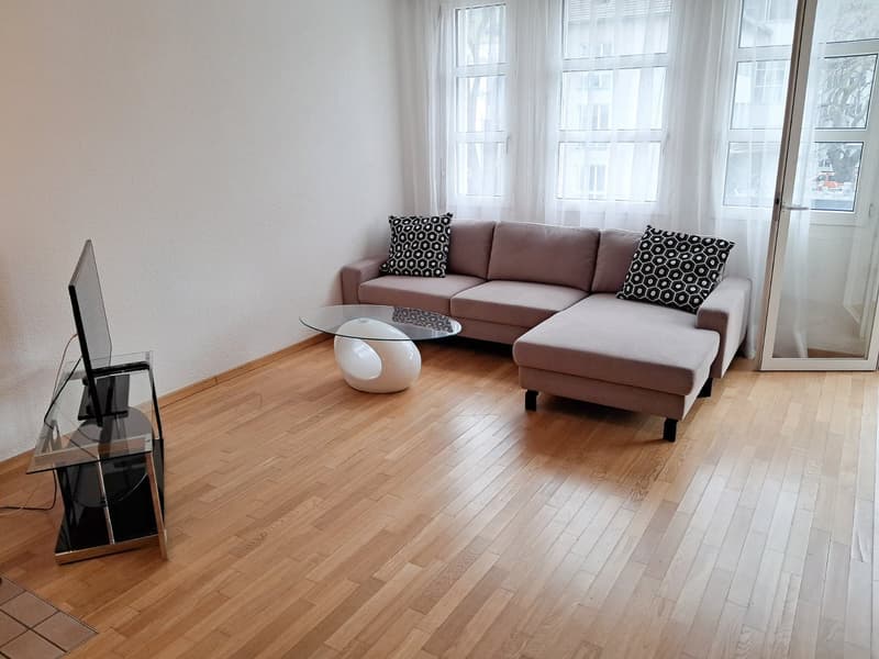 Spacious living room with hardwood floors, large windows, a beige sectional sofa with patterned throw pillows, and a glass coffee table. The room has a modern and minimalist design.