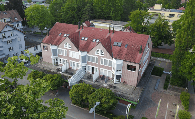 Multi-story building with red tiled roofs, white walls, and arched windows. The building appears to be surrounded by trees and greenery, with a paved area in front.