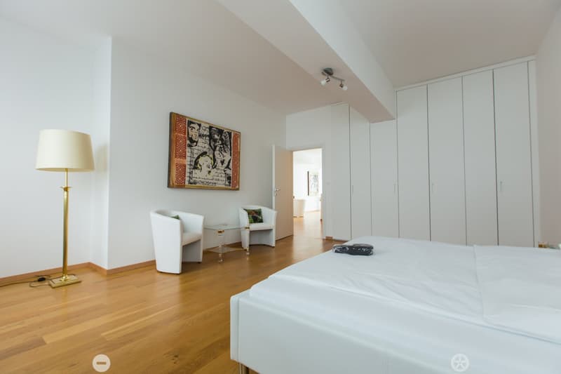 Spacious bedroom with hardwood flooring, white walls, large windows, built-in closets, and a seating area with a chair and side table. The room also features a decorative wall art piece.