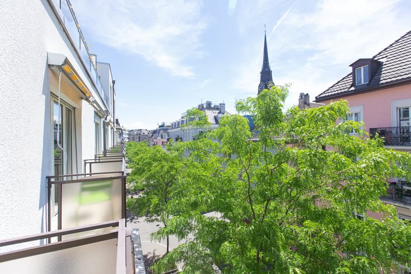 The image shows a view of a city street with a church steeple visible in the background. The foreground features a balcony or terrace with lush greenery and trees. The buildings along the street have a mix of architectural styles and colors, creating a ch