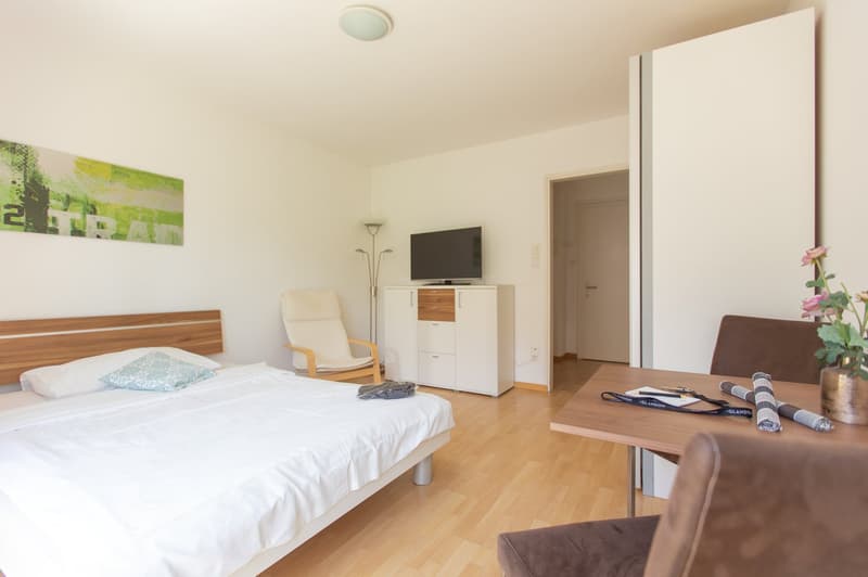 Spacious bedroom with hardwood flooring, white walls, a large bed with white bedding, a wooden headboard, a TV stand with a flat-screen TV, a comfortable armchair, and a small table with some decor items.