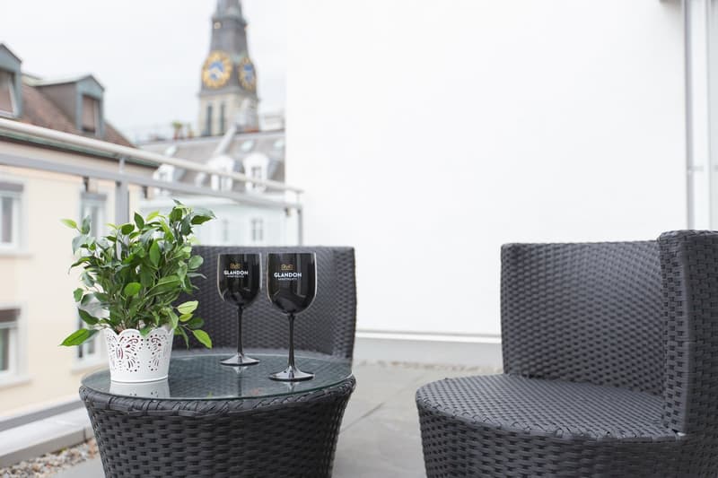 glass table, two wine glasses, green plant, two wicker chairs, glass railing