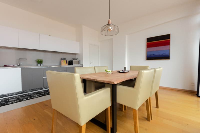 The image shows an open-concept kitchen and dining area. The kitchen features white cabinets and a stainless steel appliance, likely a dishwasher. The dining area has a wooden table with four beige upholstered chairs. A decorative pendant light fixture ha