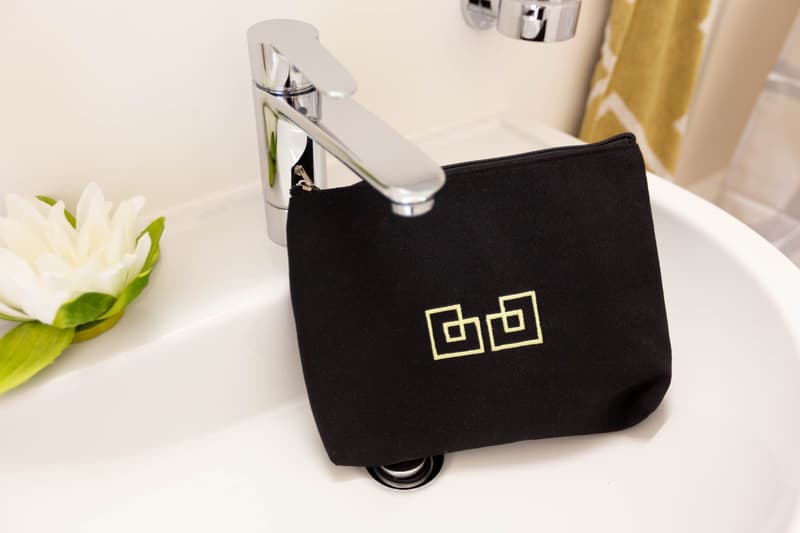 Black cosmetic bag with a white logo, placed next to a chrome faucet and a white flower on a white surface