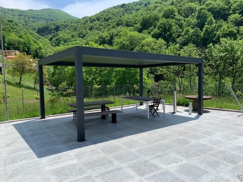 covered seating area with tables and chairs, paved floor, metal frame structure, fenced area with greenery in the background