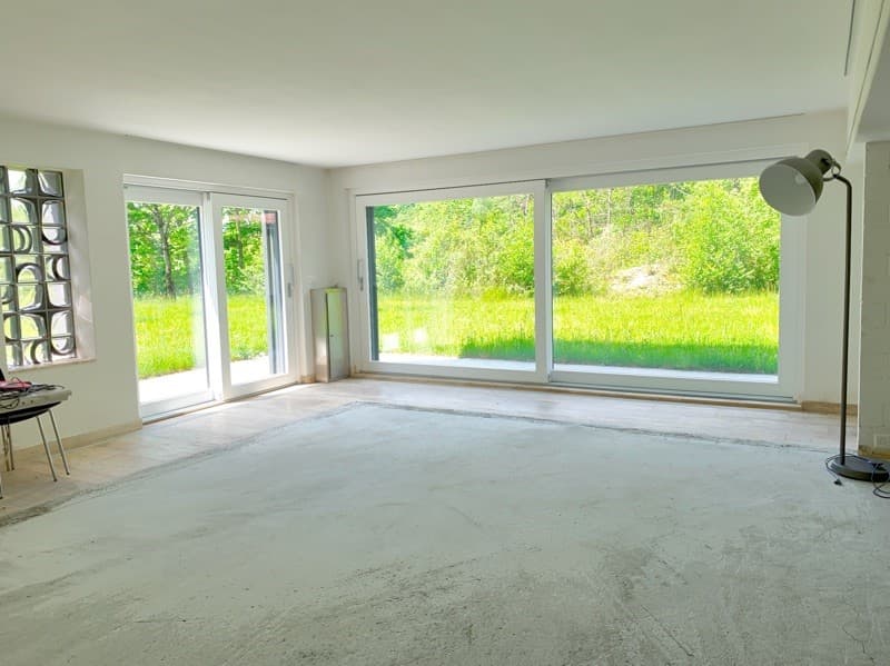 empty room with white walls and windows, grey carpet, standing lamp, table by the window