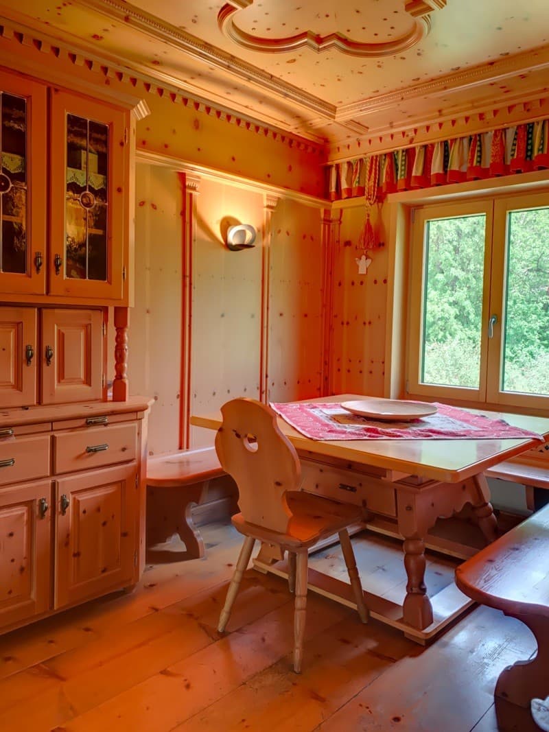 Dine room with large wooden dining table, chairs, and benches, a wood cabinet with glass doors, and windows with view to the outside.