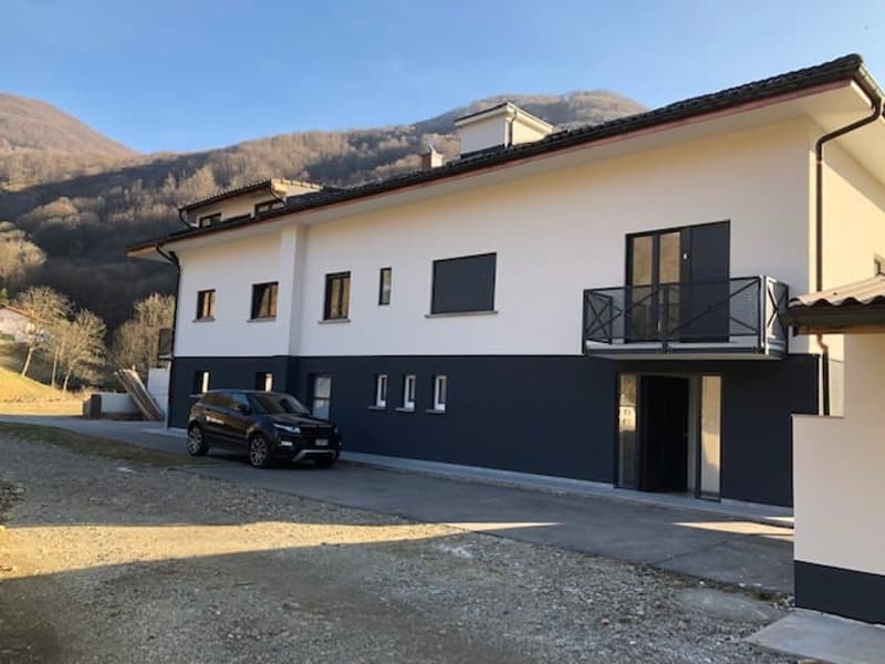 Modern two-story house with black and white exterior. It features a balcony with a railing, a black car parked in front, a sloping driveway, and a picturesque view of a mountain range in the background.