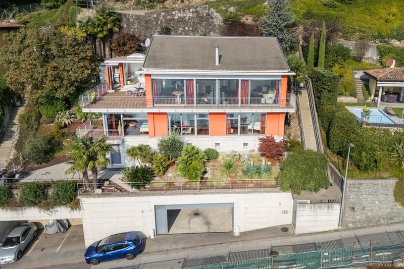 two-story house with glass windows, balcony, garage, swimming pool, cars parked, plants, stones on the ground, surrounded by plants and trees
