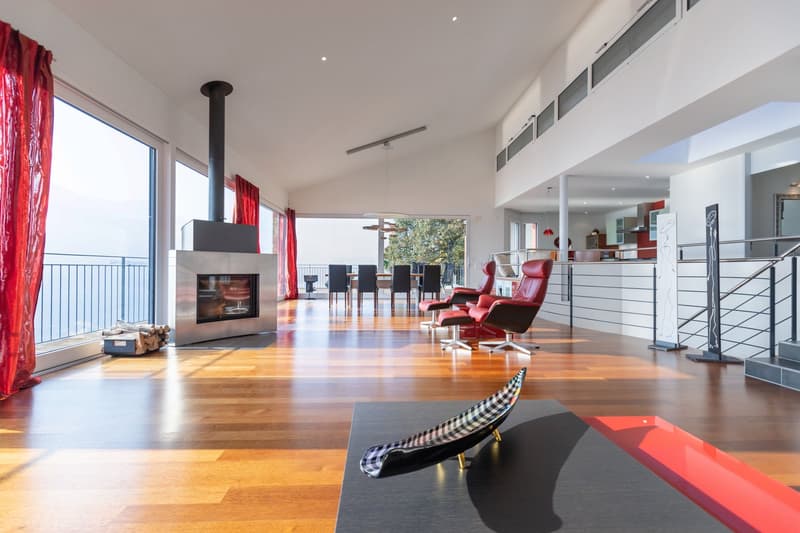 Modern living room with hardwood floors, white walls, large windows, fireplace, and balcony. It has a dining area with a round dining table and red chairs. Additionally, there is a staircase and a balcony with a railing.