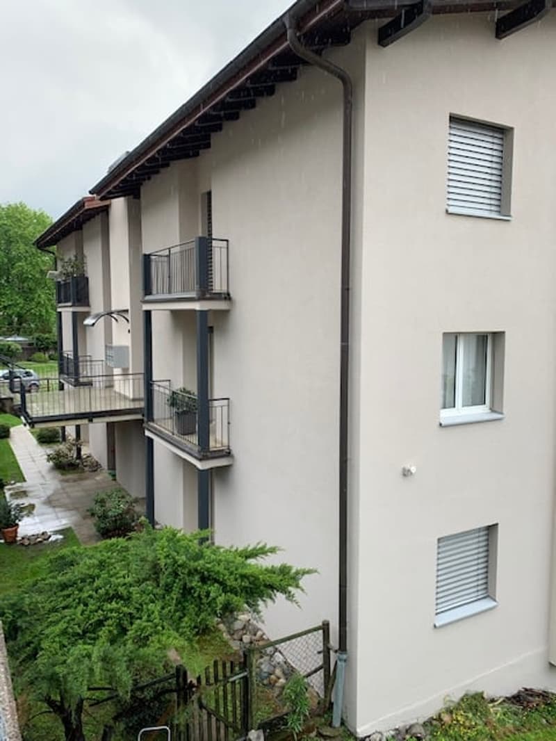 3 story apartment, beige painted, windows with blinds, balconies, plants, paved pathway, greenery