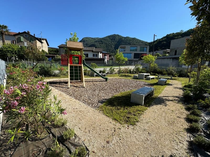 A small, fenced garden with a gravel playground featuring a slide and climbing frame, surrounded by plants and trees, with benches and green grass.