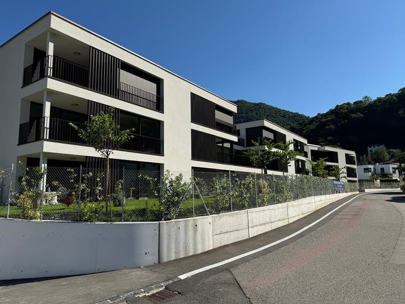 Multi-story apartment building, white exterior, black balconies, fenced garden, concrete barrier, road, greenery