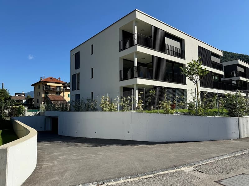 A white modern building with multiple floors, balconies, and a parking entrance, surrounded by greenery and trees, located in a peaceful area.