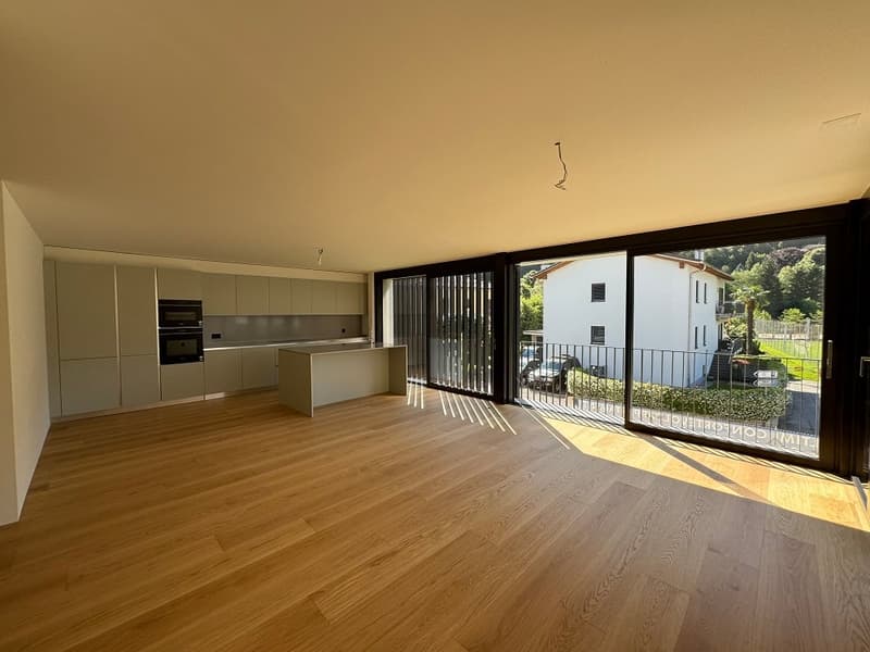 Spacious open-plan living area with hardwood flooring, modern kitchen with built-in appliances, and large sliding glass doors leading to a balcony or terrace with a view of the surrounding greenery.