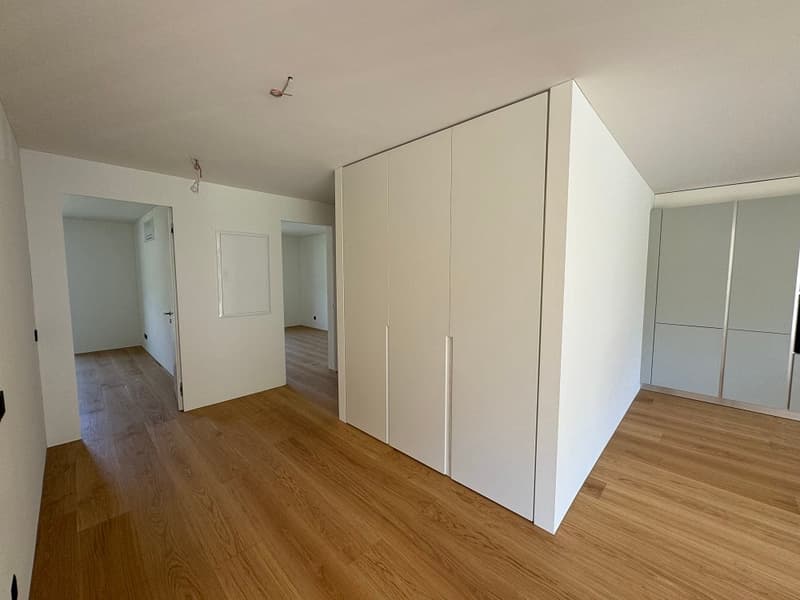 Spacious bedroom with white walls, hardwood flooring, and a large built-in wardrobe system. The room appears to have ample natural light and a clean, minimalist design.