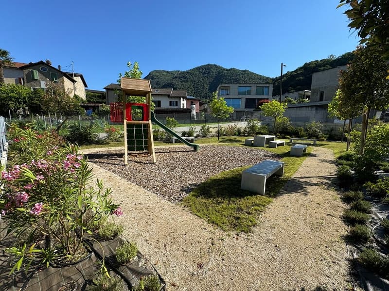 The image shows a well-maintained outdoor community space with a playground, benches, and lush greenery. The space features a slide and other play equipment for children, surrounded by a gravel path and landscaped gardens. The buildings in the background
