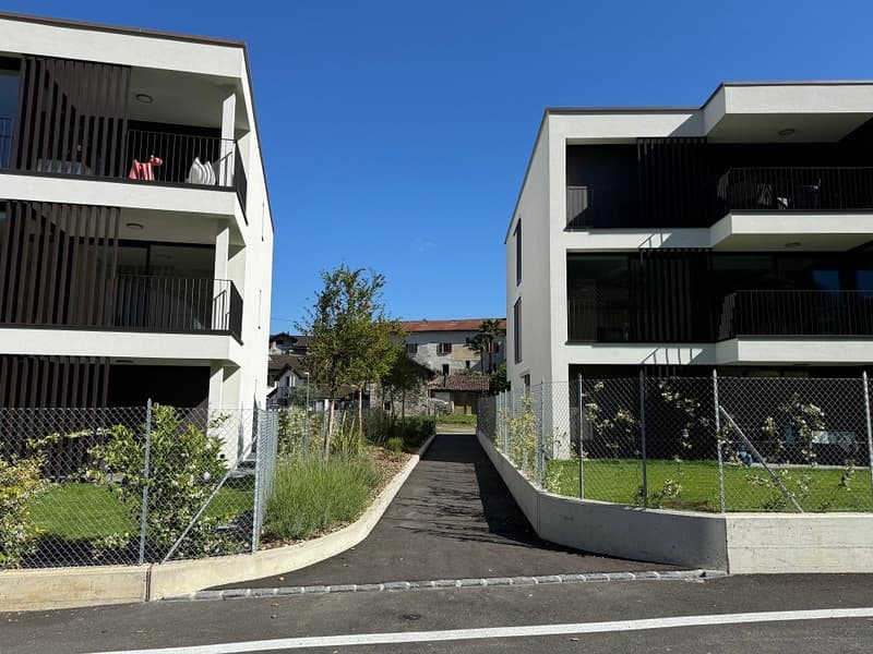 Two multi-story apartment buildings with balconies, surrounded by greenery and a paved walkway.