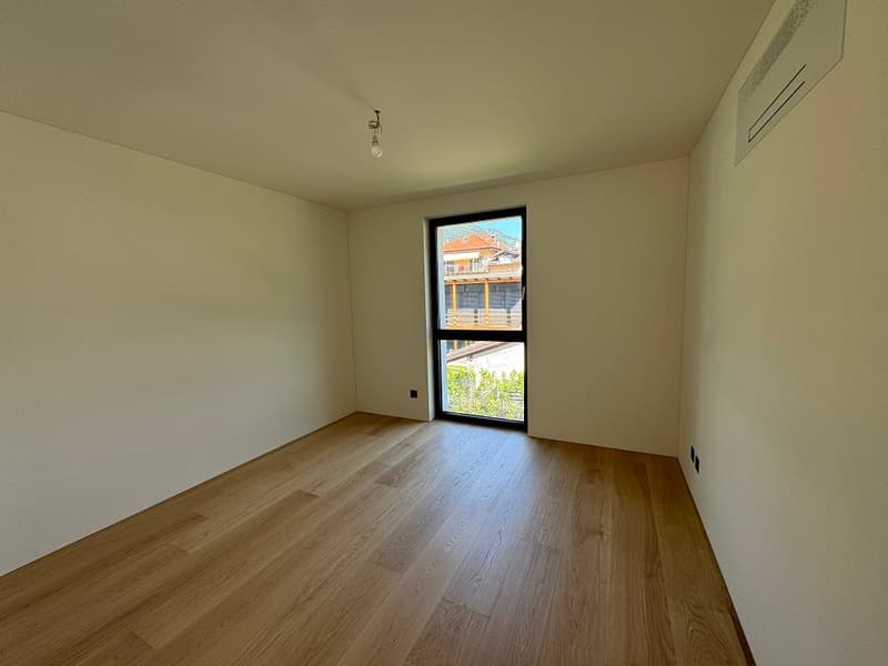 Spacious bedroom with hardwood flooring, white walls, and a large window providing natural light and a view of the exterior.