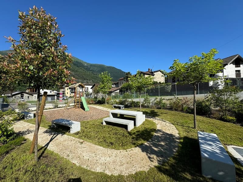 Outdoor playground with benches, gravel path, and green grass surrounded by residential buildings and trees