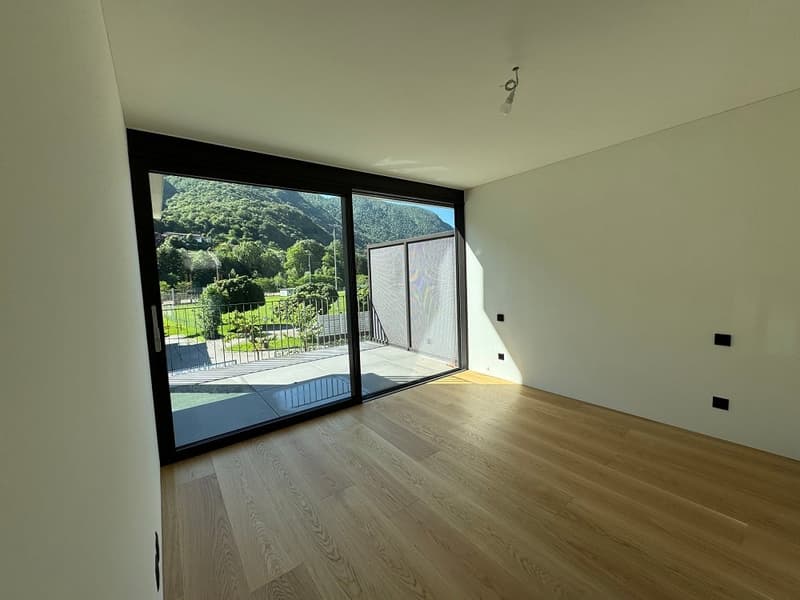 Spacious living room with large windows overlooking a garden, hardwood flooring, and a sliding glass door leading to a balcony or terrace.