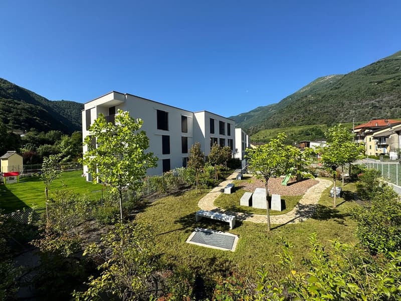 Multi-story apartment building with a modern, minimalist design surrounded by a well-maintained garden and landscaping. The building has large windows and balconies overlooking the mountainous terrain in the background.