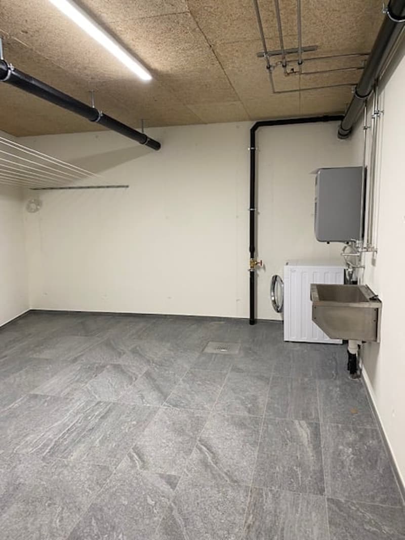 This image shows a utility room or laundry room with a washing machine, exposed pipes and ductwork on the ceiling, and a tiled floor.