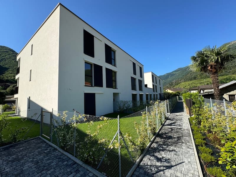 A modern, multi-story apartment building with a gray exterior and large windows. The building is surrounded by a grassy area with a paved walkway leading to the entrance. There is a palm tree visible in the background, indicating a warm, tropical climate.