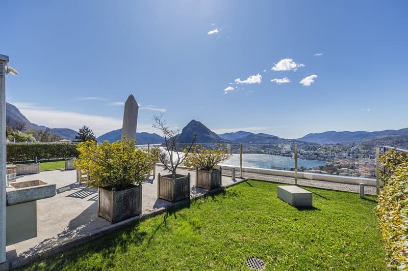 Outdoor area with a view of the mountains and the lake, green grass, wooden planters with plants, concrete benches, and a metal fence.