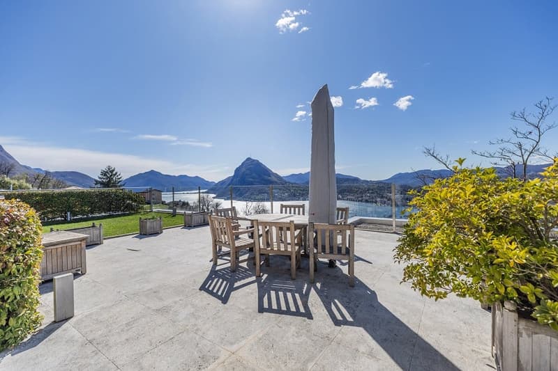 Outdoor dining area with table and chairs, umbrella, surrounded by plants and bushes, overlooking a lake and mountains