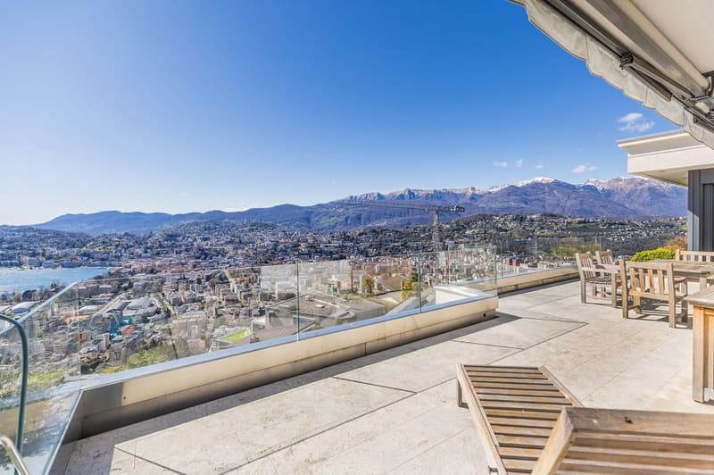 large balcony with glass railing, wooden chairs and table, view of the city and mountains