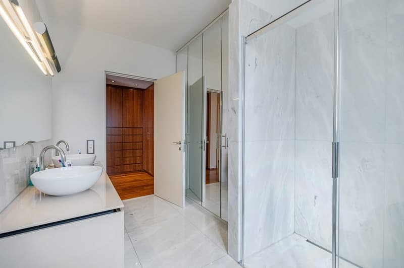 modern bathroom with white marble tiles, two sinks, glass shower enclosure, wooden door, wooden floor