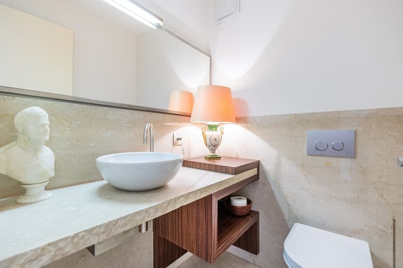 white sink, wooden cabinet, large mirror, white toilet, bust sculpture, lamps, marble wall