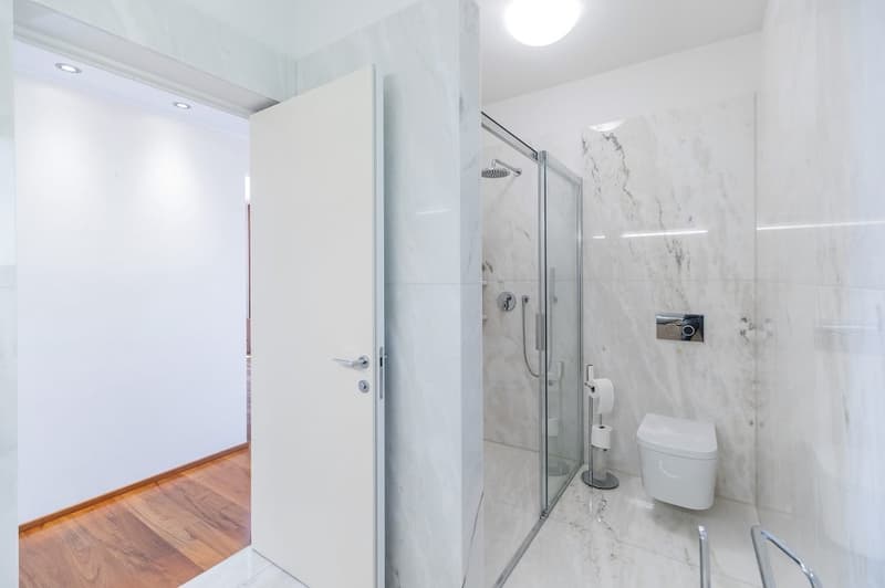 modern bathroom with white marble tiles, glass shower enclosure, white toilet, wooden floor