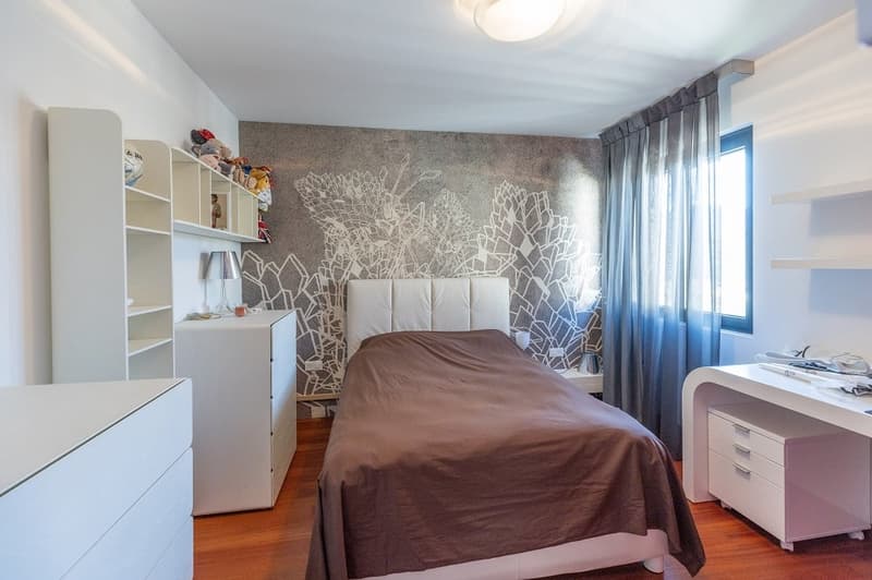 Bedroom with bed, white headboard, white shelves, white desk, brown bedspread, white walls, wooden floor, lamp, window with blue curtain