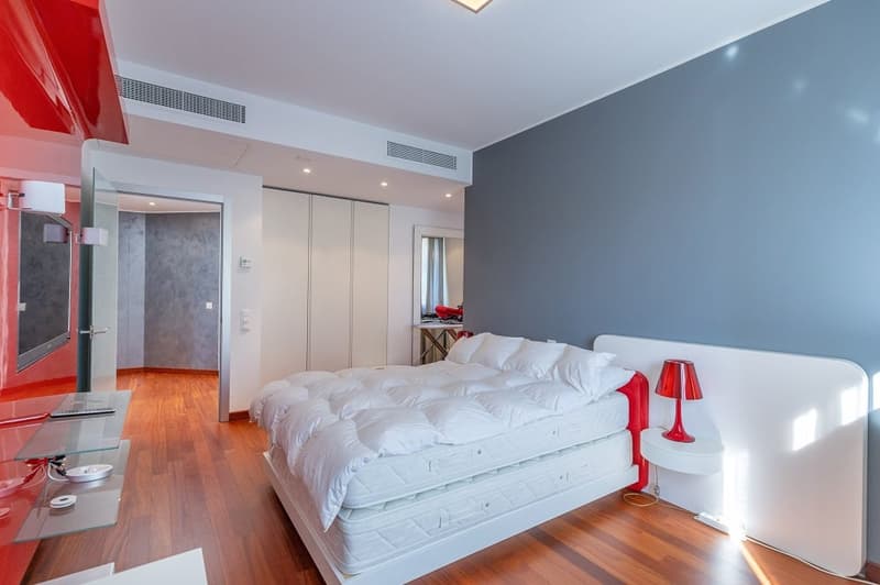 Bedroom with a large bed, white headboard, red lamp, wooden floor, gray walls, white cabinets, and a glass table with a laptop.