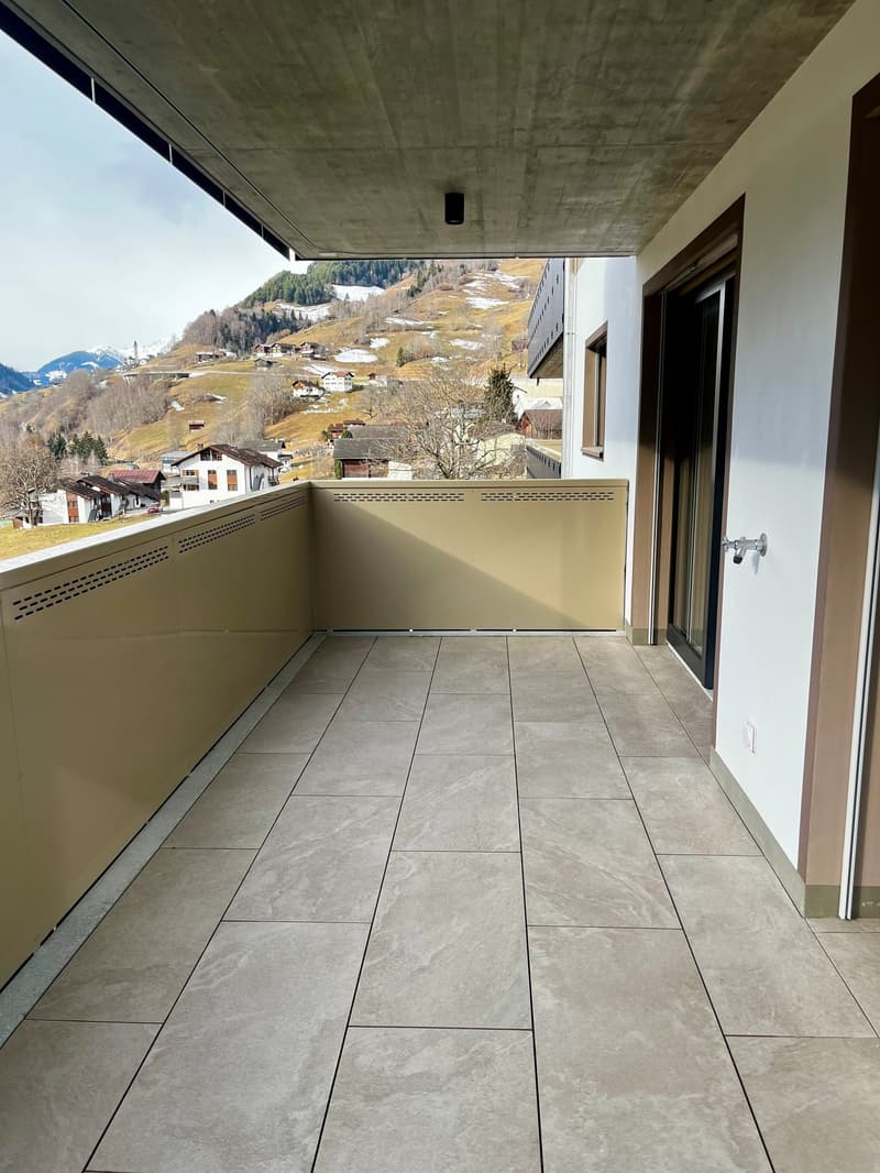 A balcony with tiled flooring and a clear view of a snowy mountain landscape. The balcony has a glass railing.
