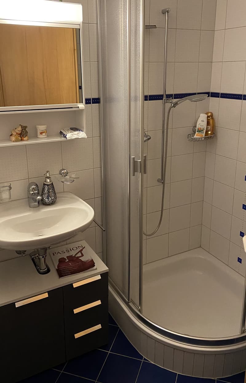 Tiled floor, white walls, silver showerhead, white sink, wooden cabinet with mirror above, shelving unit with toiletries, and a power outlet near the wall.